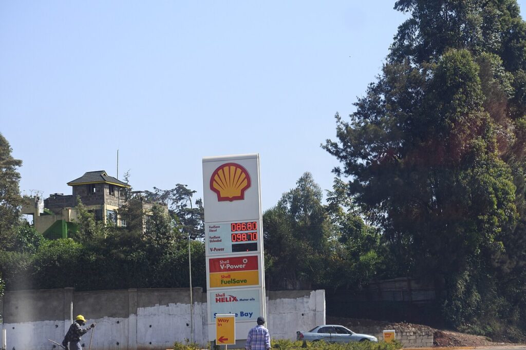 A fuel station forecourt at dusk with white cylindrical petroleum storage tanks, tall fuel pumps, and a fuel tanker truck parked beside the