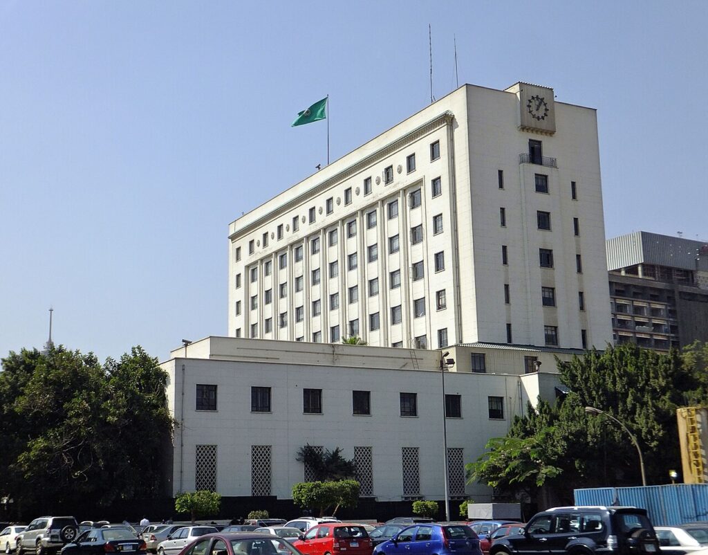 The Arab League headquarters building in Cairo showing its distinctive curved facade and columned entrance, with diplomats walking toward th