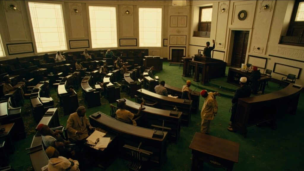 An interior view of a parliamentary debating chamber with rows of seated legislators in robes, one figure standing and gesturing at the podi