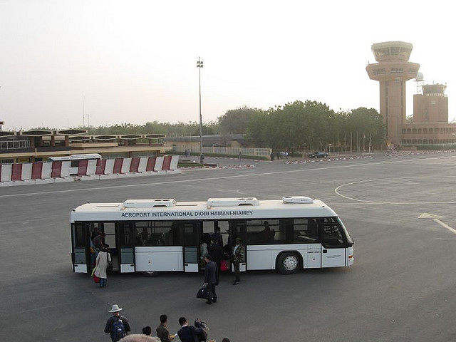 A wide exterior view of a regional airport terminal building in a flat, arid Sahelian landscape, with an empty perimeter road and distant lo