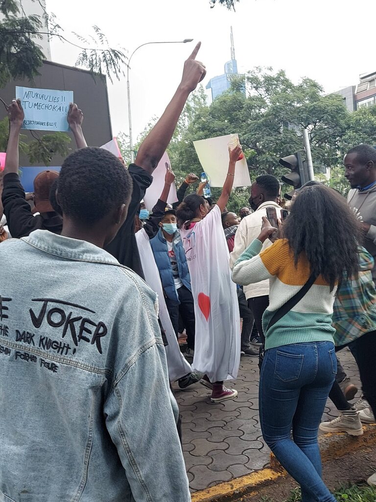 A wide street-level view of a dense crowd of young demonstrators marching down a broad city avenue, photographed from behind at mid-distance
