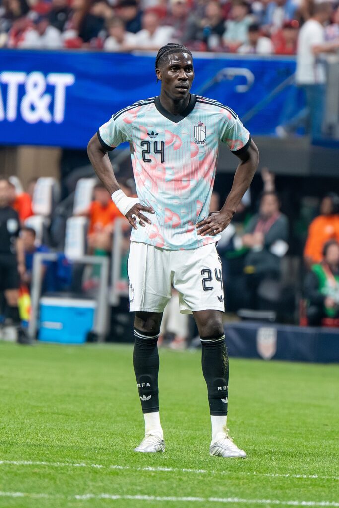 Football players in light-coloured jerseys celebrating together near the goal area inside a floodlit modern stadium at night, with a densely