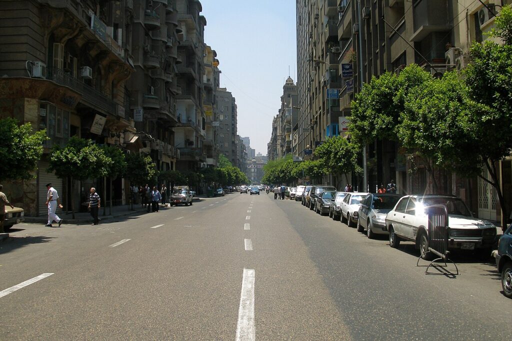 A bustling North African city avenue at dusk packed with football supporters in red and white, tricolor flags draped from balconies and cars