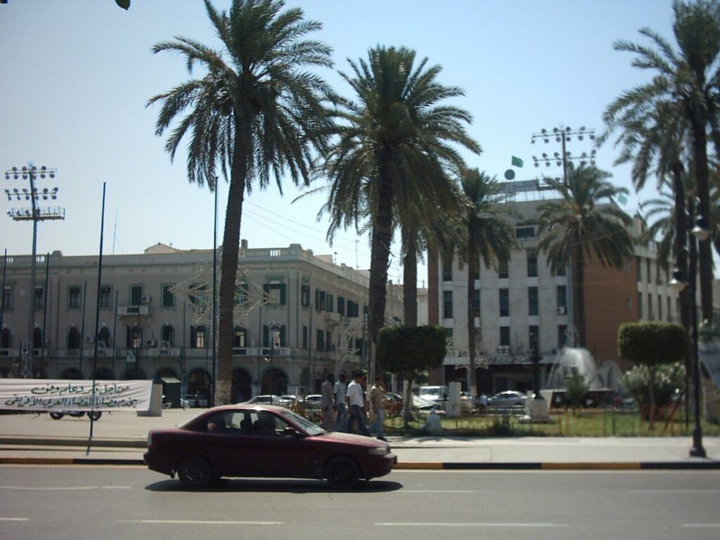 A nighttime protest on a wide Tripoli boulevard, with crowds gathered at an intersection behind makeshift barricades as small fires burn and