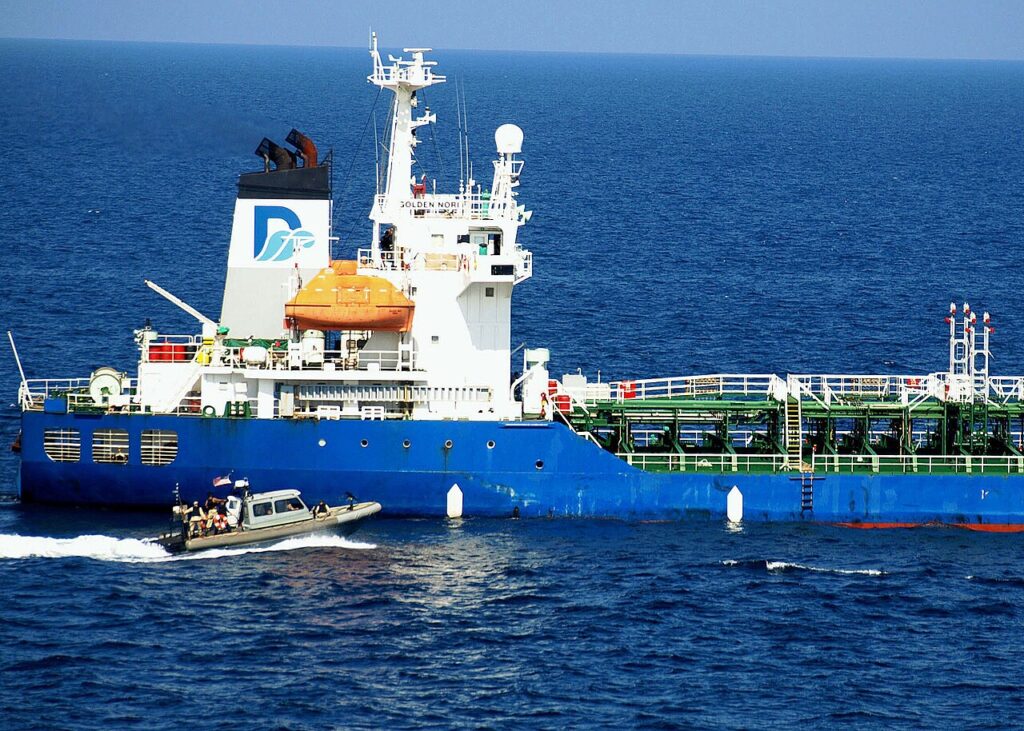 A large grey commercial oil tanker steaming across open sea at dusk with a small dark skiff approaching from the side, shot from mid-distanc