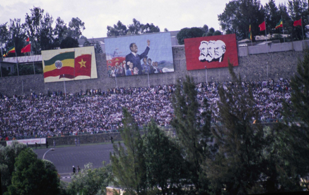 A dense crowd of demonstrators marches along a wide city avenue, uniformed observers at the edges and mid-rise buildings lining the route.