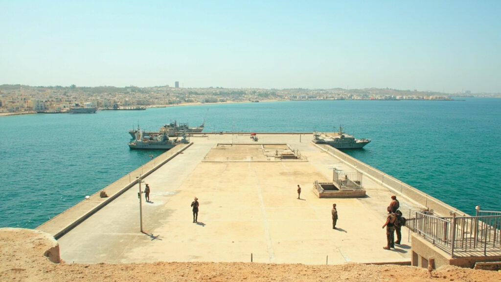 A wide view of a Mediterranean port city with an industrial harbor, cranes, and naval vessels docked along the coastline under hazy daylight