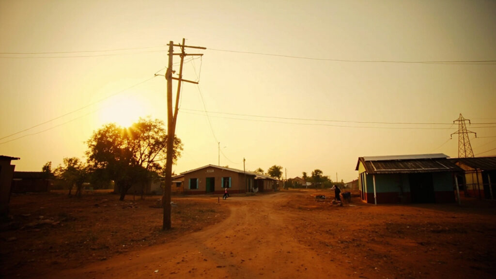 A rural African village scene showing a wooden utility pole with electrical wires and a small rooftop solar panel on a modest building.