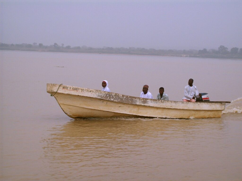 An overcrowded wooden passenger boat carrying people and cargo on a wide muddy river in Nigeria, with smaller boats visible along the tree-l