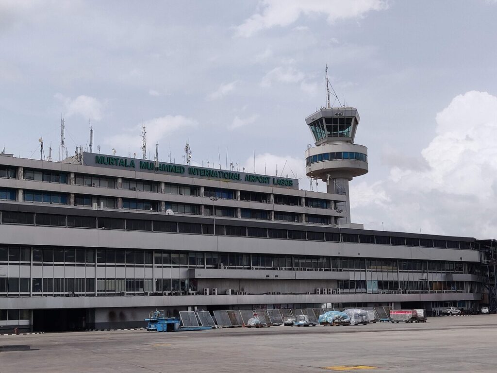 An interior view of a Nigerian airport terminal with check-in counters, passengers and airline staff under fluorescent lighting.