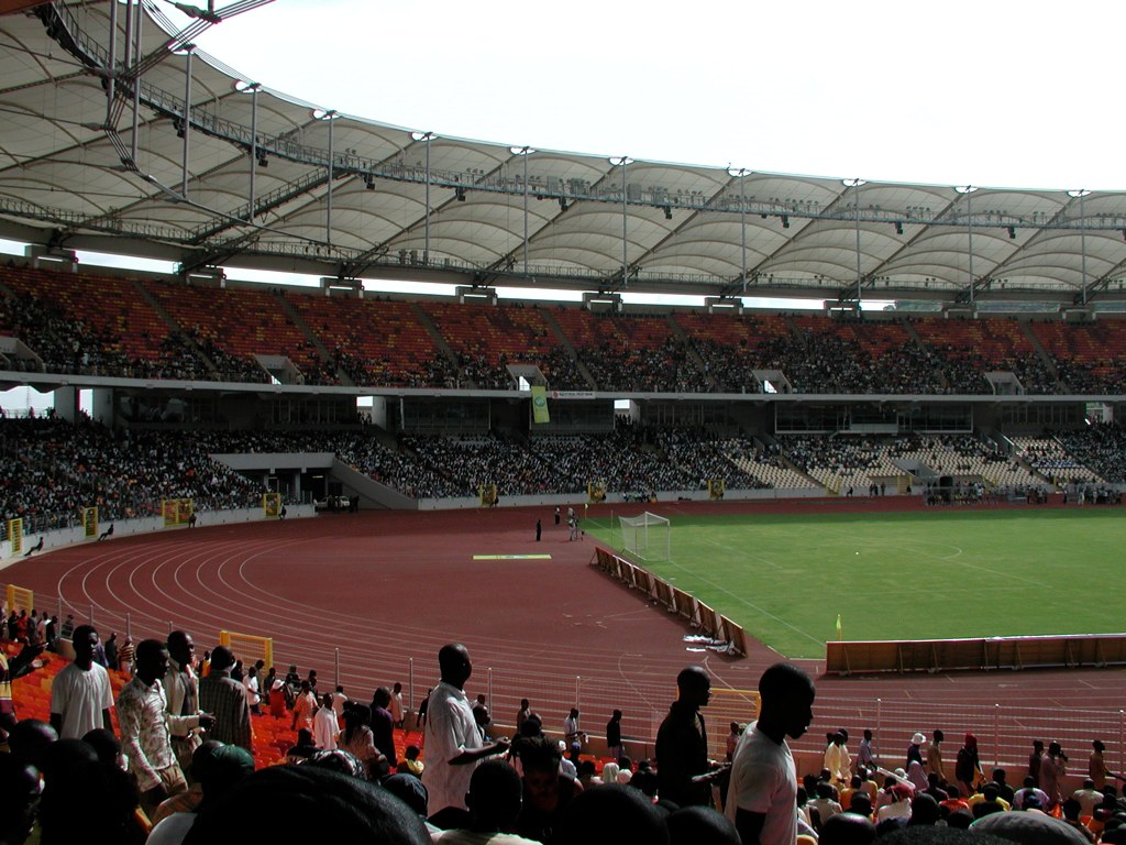 A wide view of a large football stadium at dusk with floodlights illuminating the pitch and empty tiered seating.