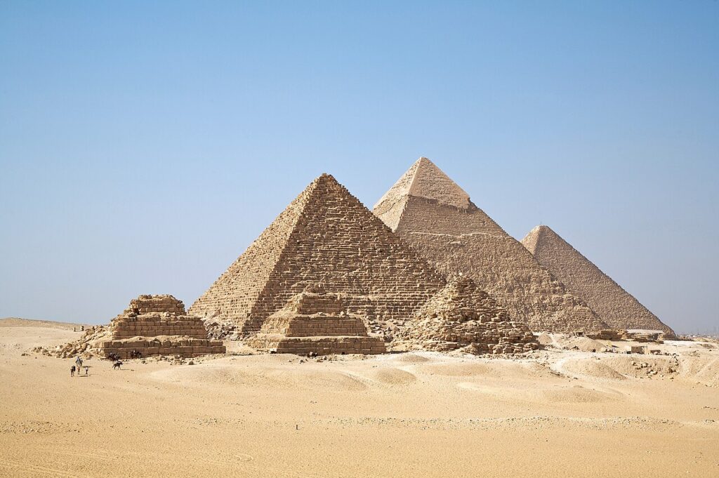 A tourist rides a camel on a sandy desert plateau with the great pyramids rising in the background under bright daylight.