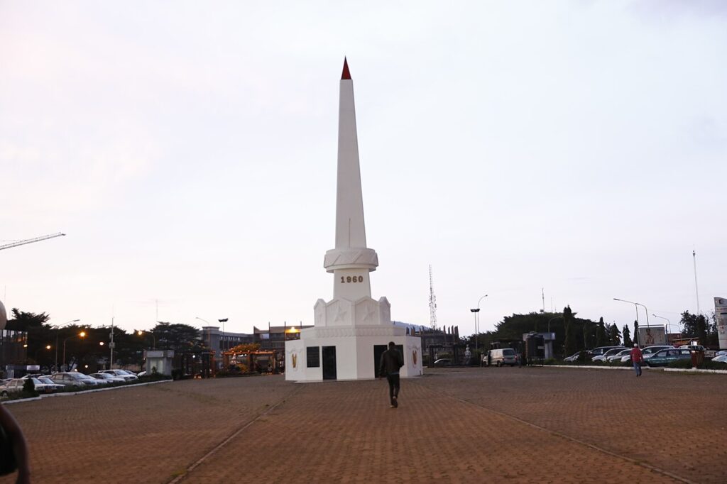 A wide view of an administrative avenue in Yaoundé with government buildings, tropical trees, pedestrians, and light traffic under bright mi