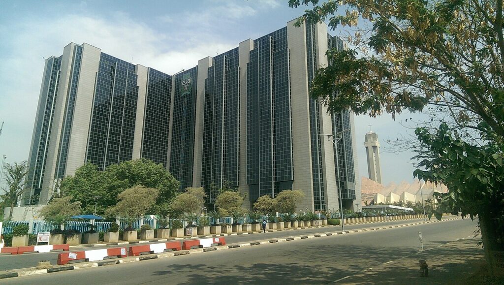 A wide exterior view of an imposing institutional government building with a columned entrance and flagpoles, set behind a manicured lawn un