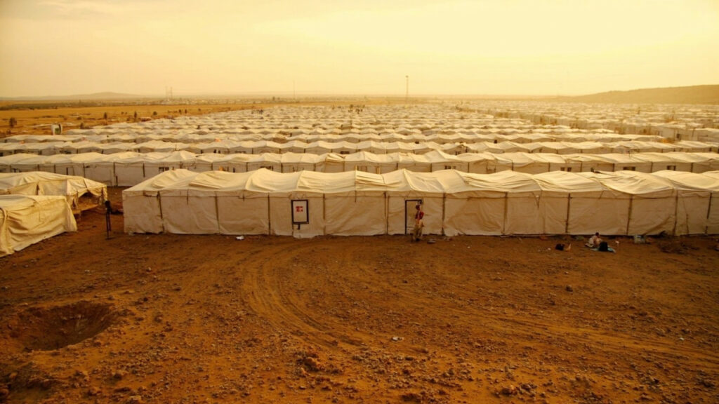 Rows of plain tents stretching across dry ground in a large displacement camp.
