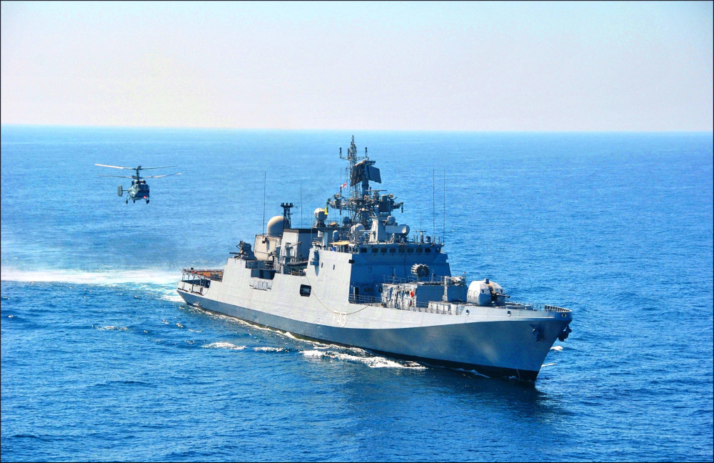 A large commercial cargo vessel underway at sea with a small armed skiff alongside it, on open blue water.