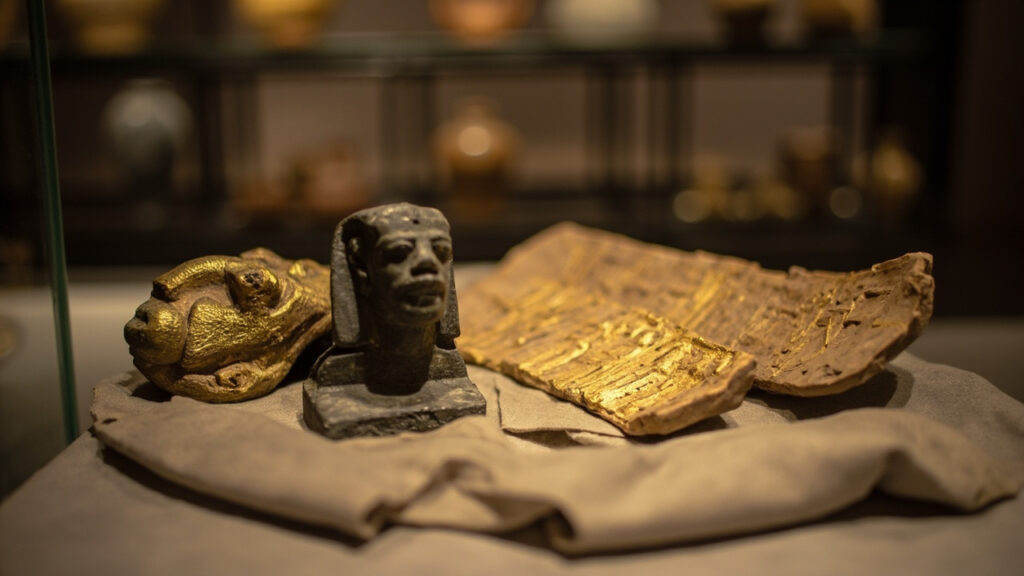 Ancient Egyptian artifacts including a gilded mummy mask fragment, a dark stone funerary statue, and a carved limestone relief panel display