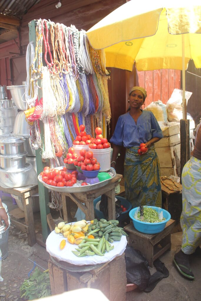 An open-air market in a tropical African city, with women traders selling produce and goods from stalls and ground-level mats under bright d
