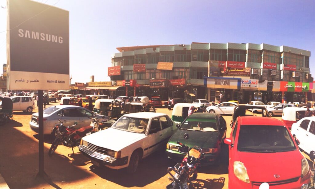 A bustling market in Omdurman with sacks of grain and produce stacked on tables, vendors behind their goods and shoppers moving between stal