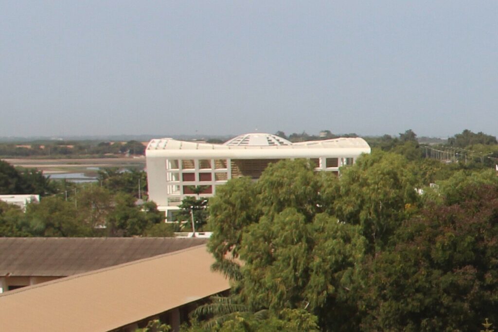 Exterior view of a colonial-era government assembly building with arched windows in a West African capital, framed by tropical trees.