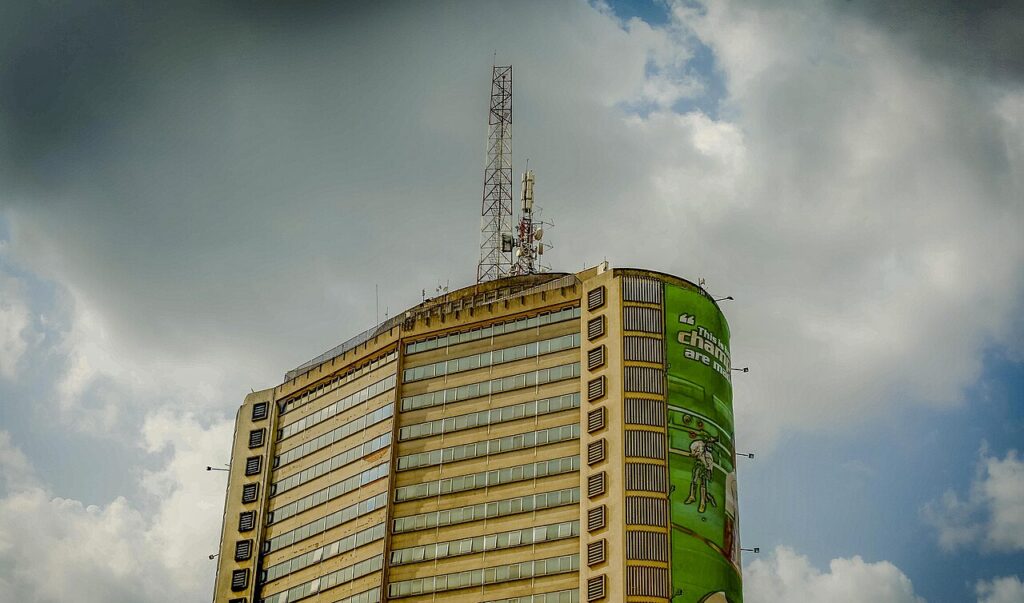 A tall modernist concrete tower in a Nigerian city with a large hourglass-shaped drum sculpture mounted on its rooftop against an amber sky.