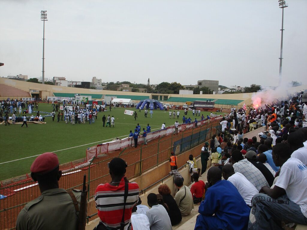 A football stadium in Dakar with a green pitch and concrete stands during daytime, with spectators near the entrance.