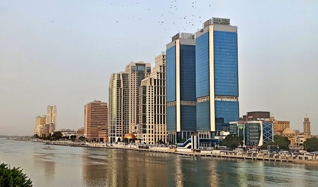 A wide photograph of a Middle Eastern capital city's skyline along a river at golden hour, showing a blend of older low-rise buildings and m