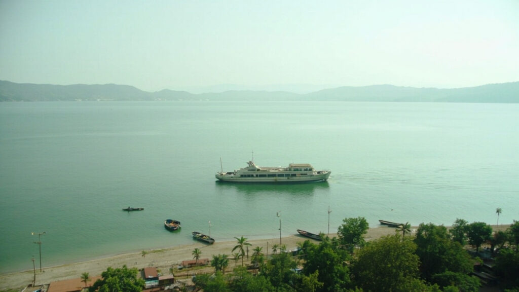 A wide view across a large calm lake with distant hills, a small passenger boat on the water, and a shoreline settlement in the foreground.
