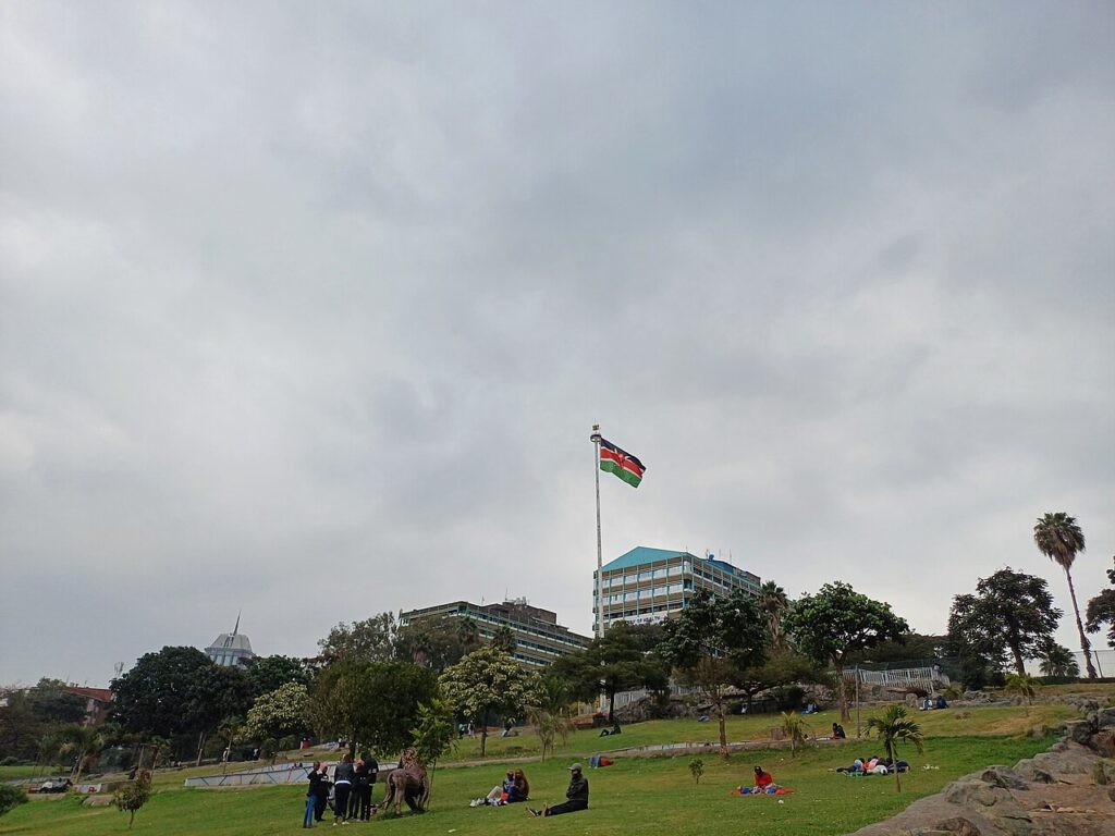 A small group of demonstrators gathered in a leafy urban park, with city buildings visible in the background under daylight.