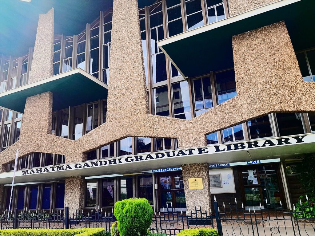 Students walking between lecture buildings on a Kenyan university campus during the day, with tropical trees and modern concrete structures