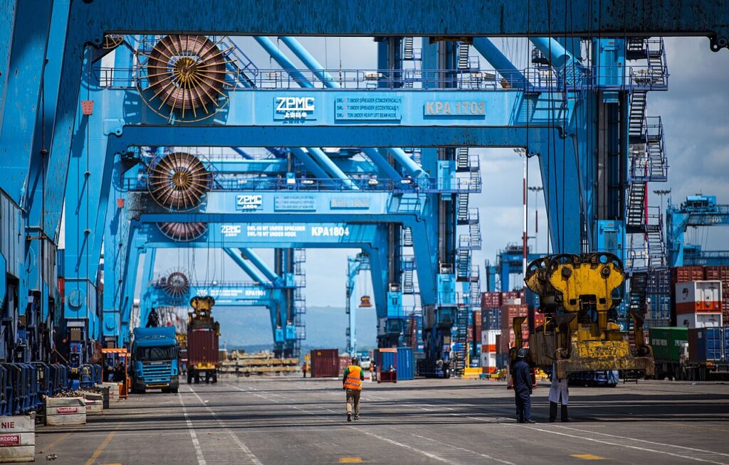 A wide view of a busy container port at dawn with gantry cranes lifting shipping containers from a docked cargo vessel.