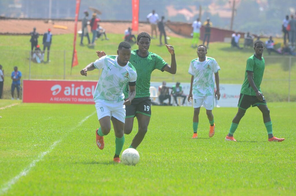 An amateur football match in progress on a grass pitch with players in colourful kits contesting possession while a small crowd watches from