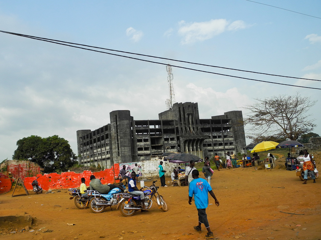 A cluster of journalists with cameras and notepads outside a low-rise government building in a tropical West African capital, photographed i