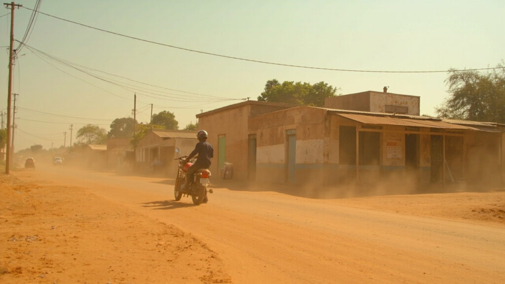 A dusty street in a small southern African town with a parked motorcycle taxi beside low-rise buildings.
