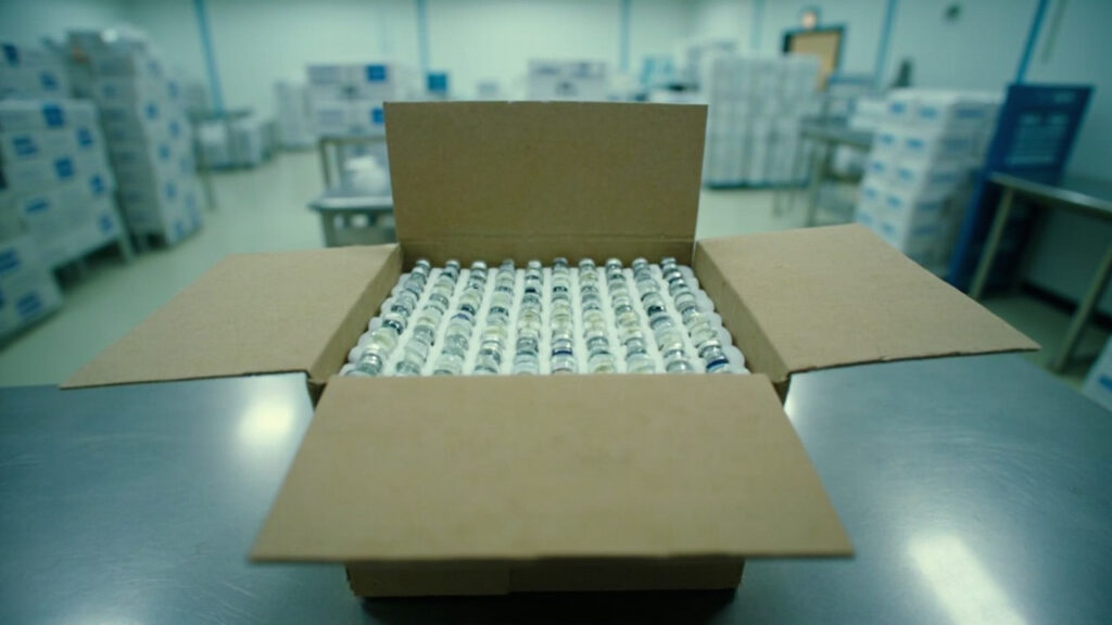 Rows of small glass vaccine vials with metal crimped caps arranged in foam inserts inside an opened cold-chain shipping box on a metal table