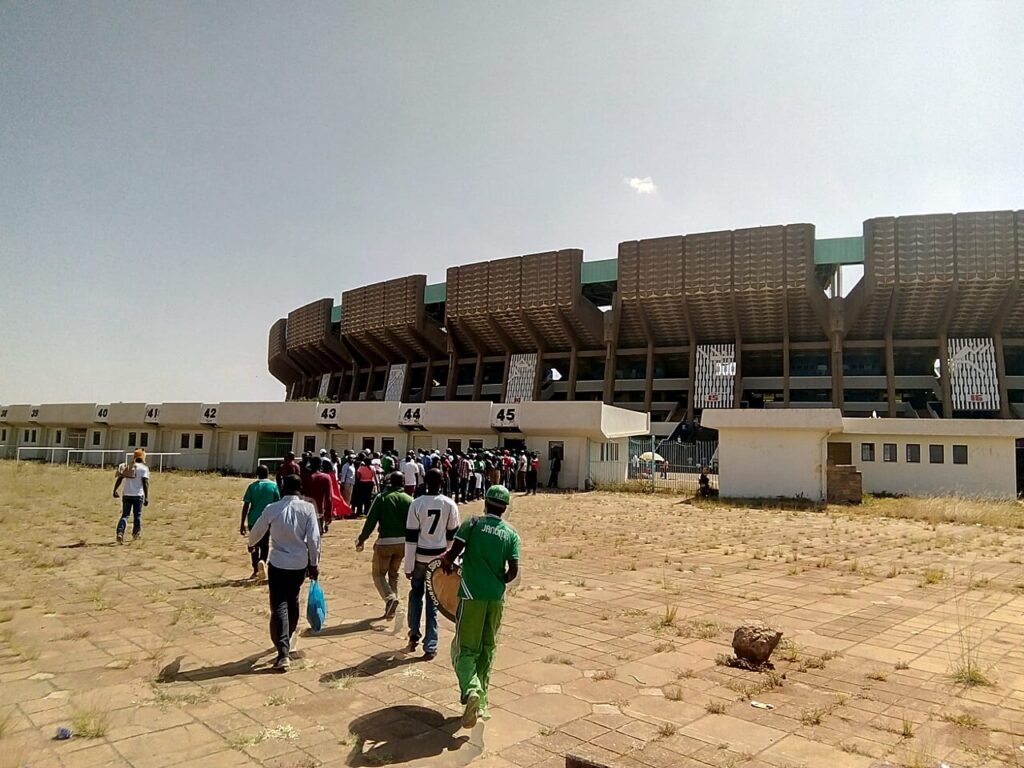 A wide exterior view of a large modern athletics stadium at golden hour, with a running track visible through the open entryways and a broad