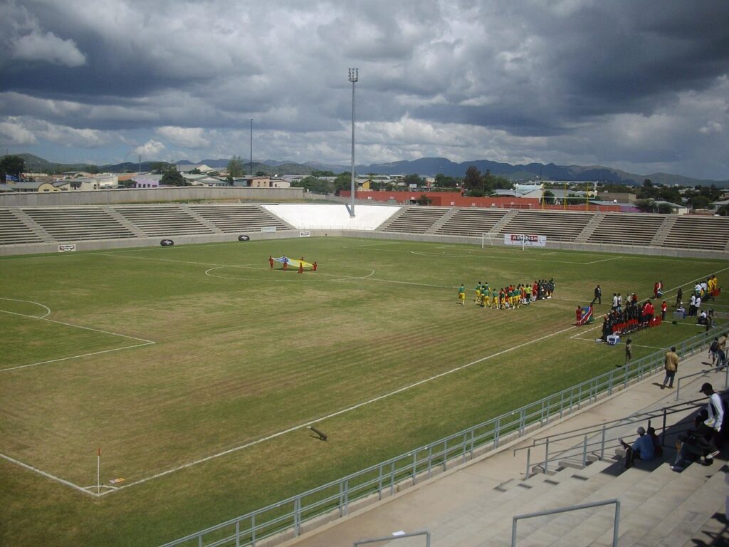 A wide exterior view of a modern football stadium in Windhoek under a clear sky, with an empty green pitch and concrete stands.