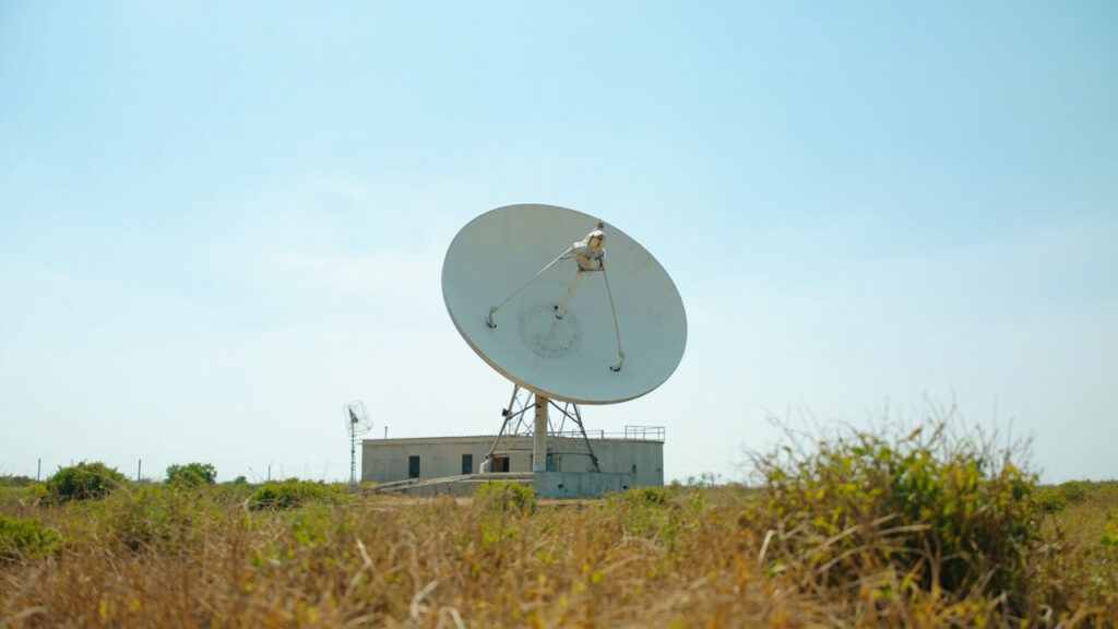 A large white parabolic satellite dish antenna at a Nigerian ground station facility under a clear blue sky.