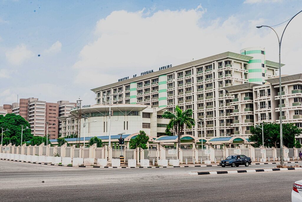 Exterior view of a large government ministry building with vehicles parked outside and security personnel at the entrance during daytime.