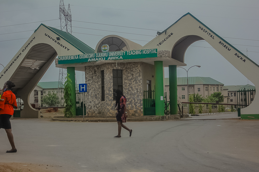 An exterior view of a Nigerian teaching hospital entrance with figures in white coats walking past a covered walkway during daytime.