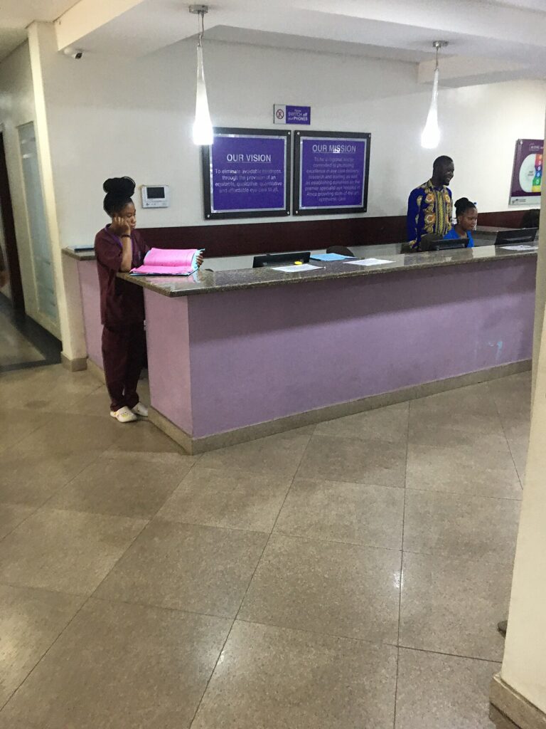 A modern hospital building in a Nigerian city, with healthcare workers visible at the entrance and patients nearby.
