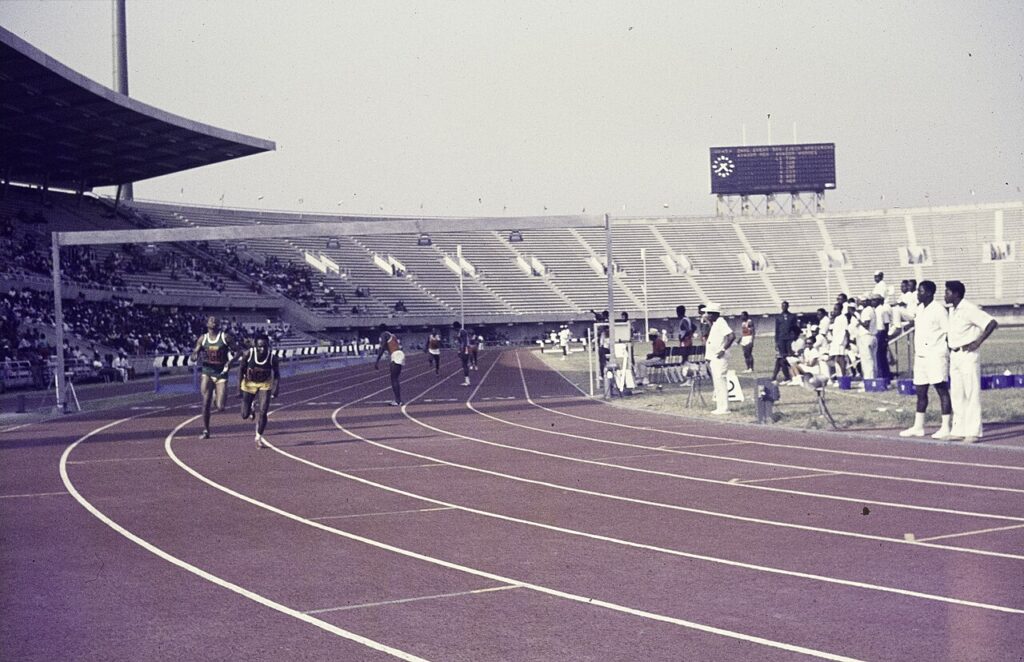 A young female athlete in a Nigerian team kit celebrates on a track with arms raised after winning a race.