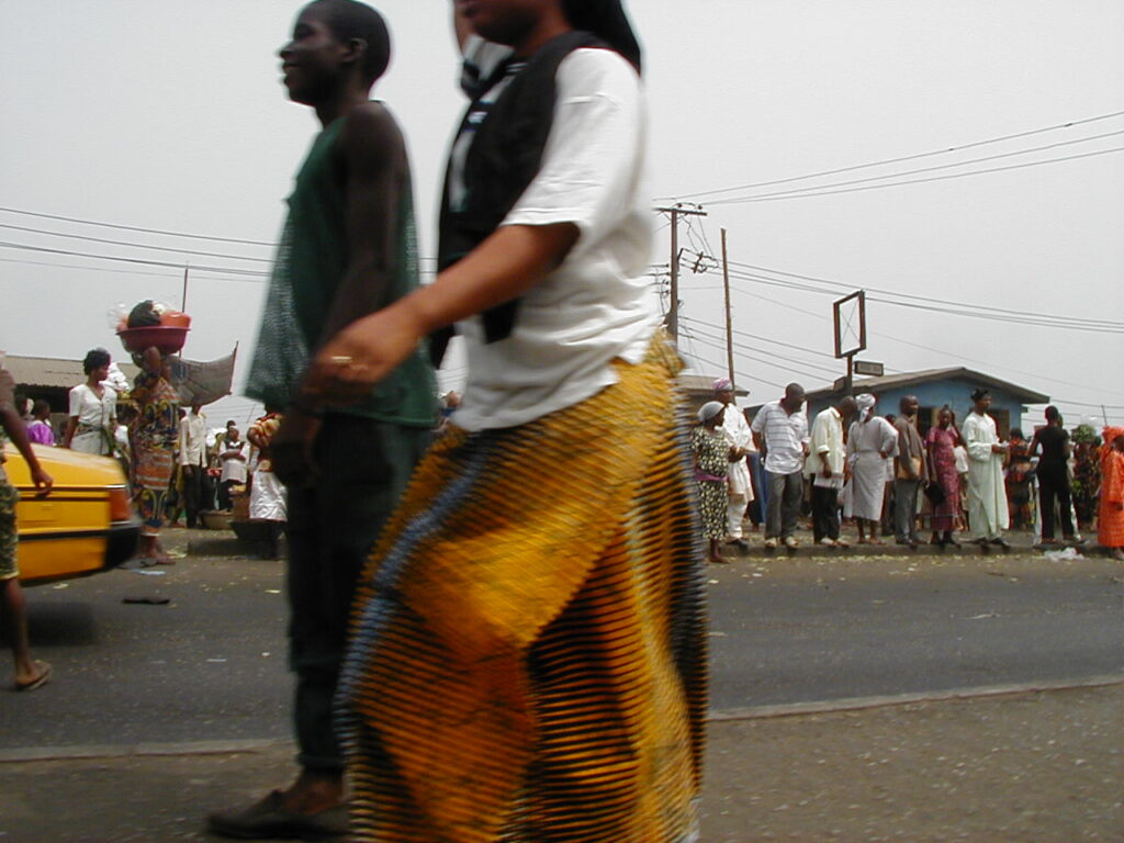 A busy West African urban street during daylight, with pedestrians, vehicles and roadside activity near a checkpoint.