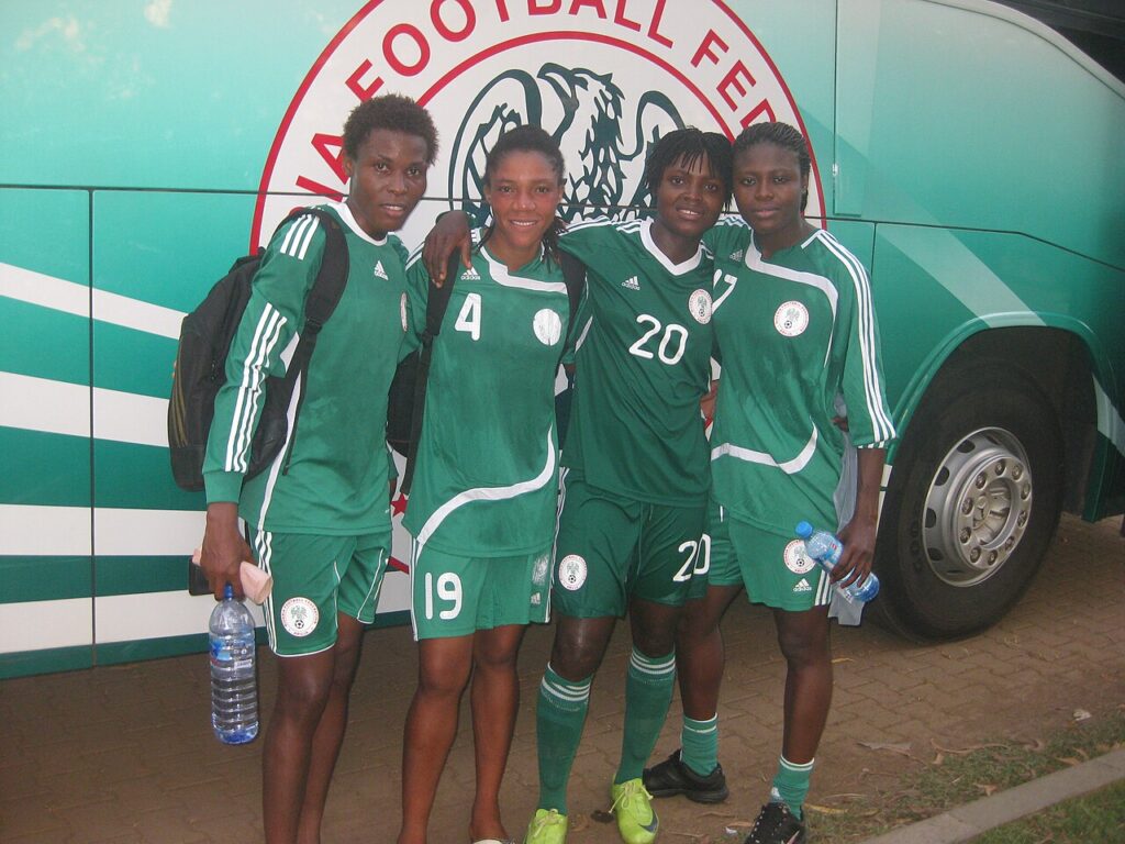Nigeria's women's football team in blue and white kits lined up on a pitch before a match, with players and a stadium in the background.