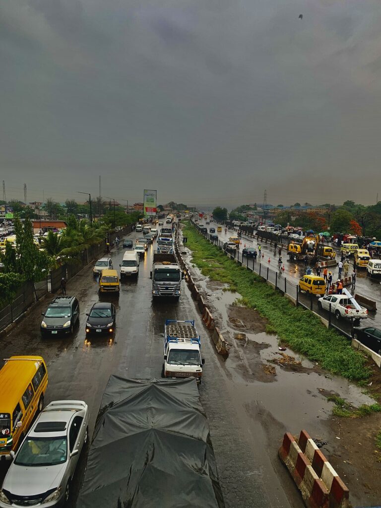 A congested urban road filled with yellow minibus taxis, motorbikes and cars under hazy skies, framed by low-rise buildings and overhead pow