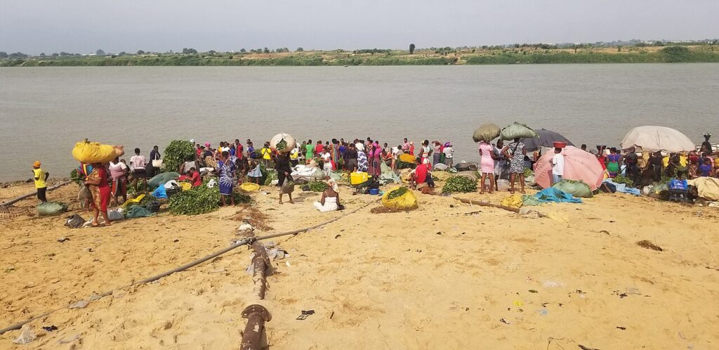 A crowded open-air market in a Nigerian riverside city, with traders, goods and wooden stalls under large awnings.