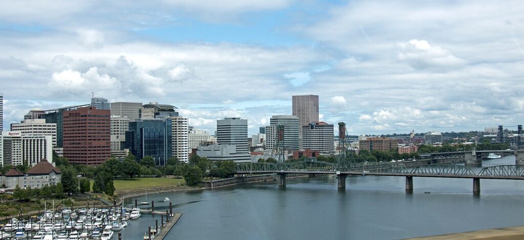 Cyclists riding across a steel-truss bridge in an American downtown with a river and skyline in the background.