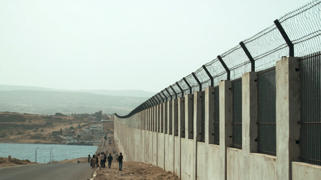 Border fence at the frontier between the Spanish enclave of Ceuta and Morocco with figures near the perimeter.