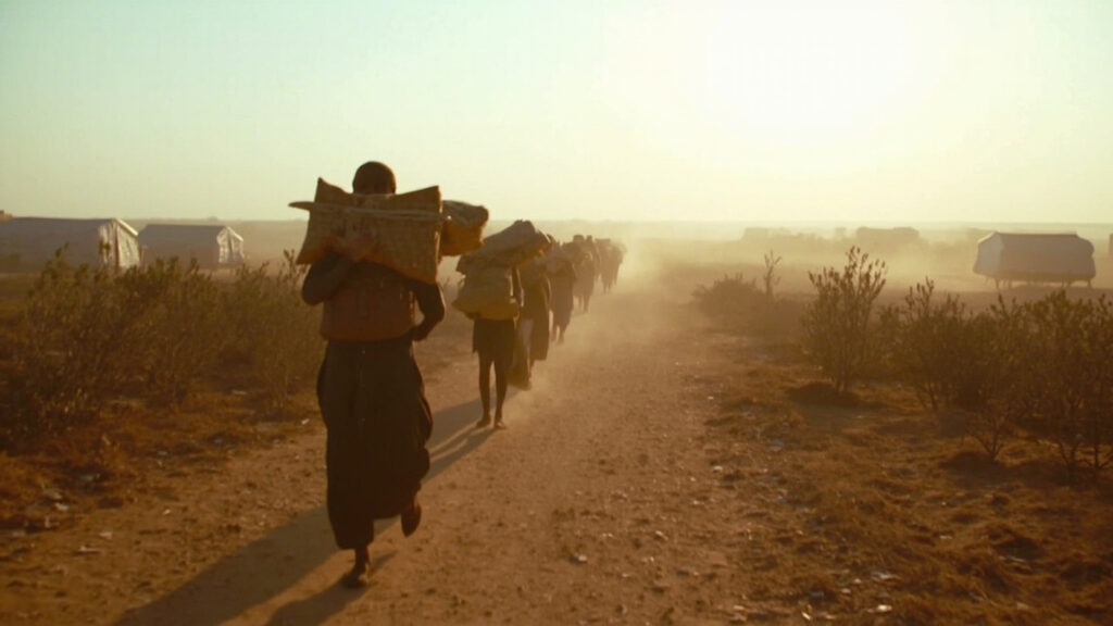 A dusty border crossing with displaced people carrying belongings walking toward a distant reception area, bundles and bags on the ground.