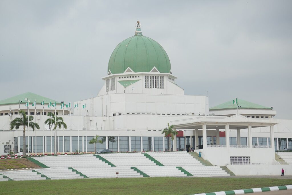 Wide exterior view of Nigeria's National Assembly complex in Abuja, framed by tropical trees and a paved plaza under bright daylight.